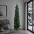 A tall artificial Christmas tree in a pine green color stands on a black metal stand in a modern living room setting with a gray sofa, framed wall art, and a large window showing a snowy outdoor view.